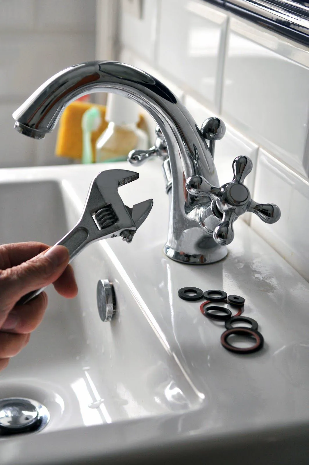 Hand holding an adjustable wrench beside a chrome basin faucet, with replacement washers laid out on the sink