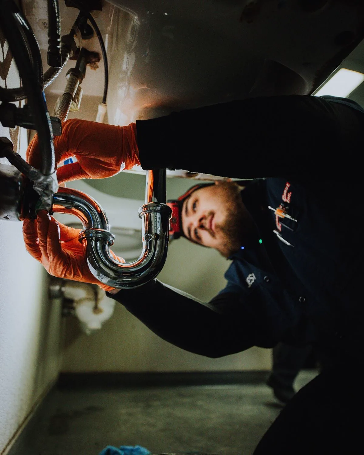 Plumber in gloves tightening a chrome P-trap beneath a sink, lit from inside the cabinet